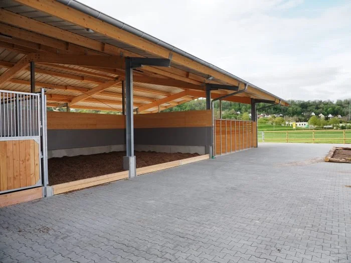 Offene Stallungen für Pferde mit Holz und Metall, im Freien auf gepflastertem Boden, mit ländlicher Umgebung im Hintergrund.