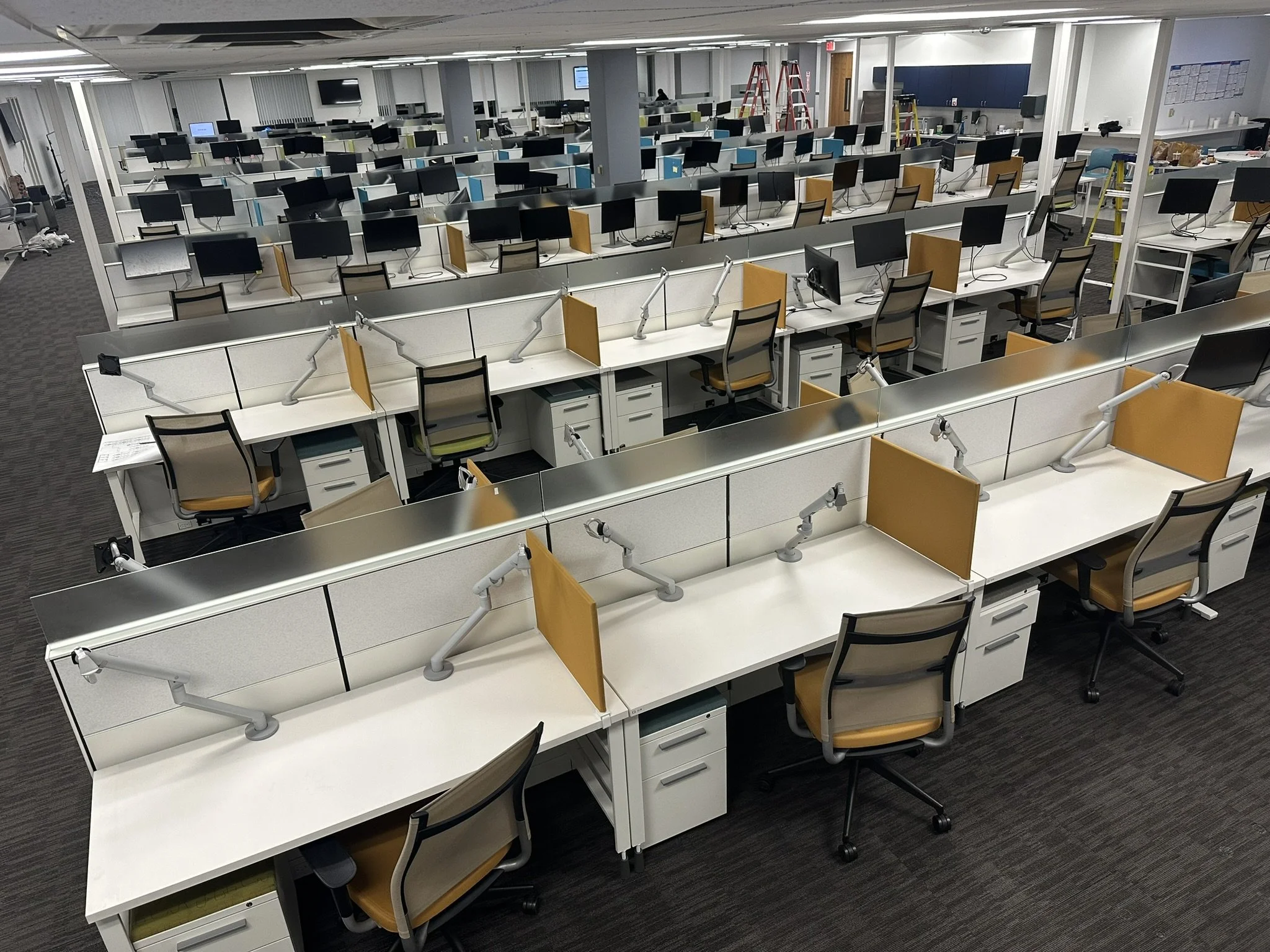 Empty office space with rows of white desks, each with monitors, chairs, and adjustable lamps. Ladders and cleaning supplies indicate active office installation, workspace setup, commercial cleaning, maintenance, and post-construction cleanup service