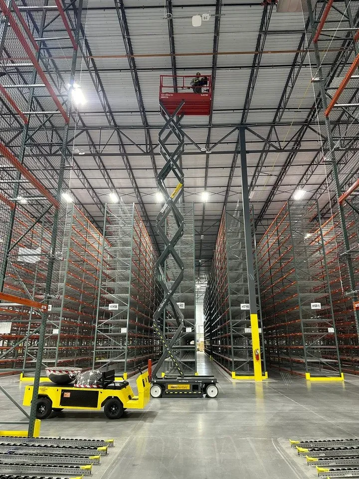 A worker on a red scissor lift working on the warehouse ceiling, with tall empty steel shelving units on either side.