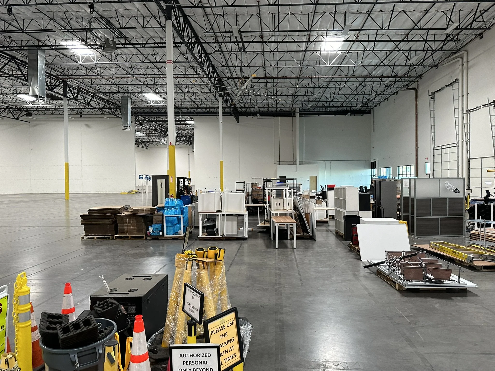 Double metal doors with small rectangular windows at the top, located in an indoor hallway with gray walls and carpeted floor. To the right is a fire extinguisher mounted on the wall, and a black stand with a fire alarm or speaker. On the walls are posters, one with a flower and the other with inspirational text and a landscape.