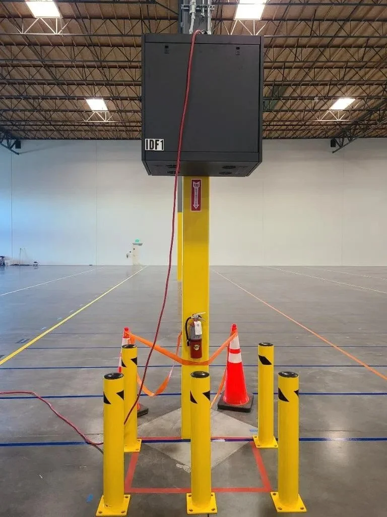 Large black speaker mounted on a yellow pole inside an empty warehouse or gym, surrounded by orange traffic cones and yellow safety posts, with a fire extinguisher attached to the pole.