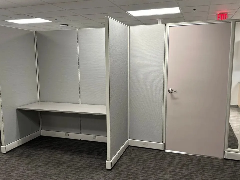 Empty office cubicle with a white desk and electrical outlets, adjacent to a closed door, in an office setting.