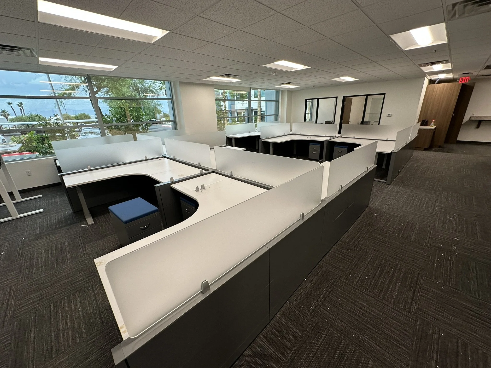 Empty office space with rows of white desks, large windows overlooking trees and parked cars, and a doorway with a red exit sign. Highlights professional office installation, workspace setup, furniture assembly, and modern commercial build-out in pro