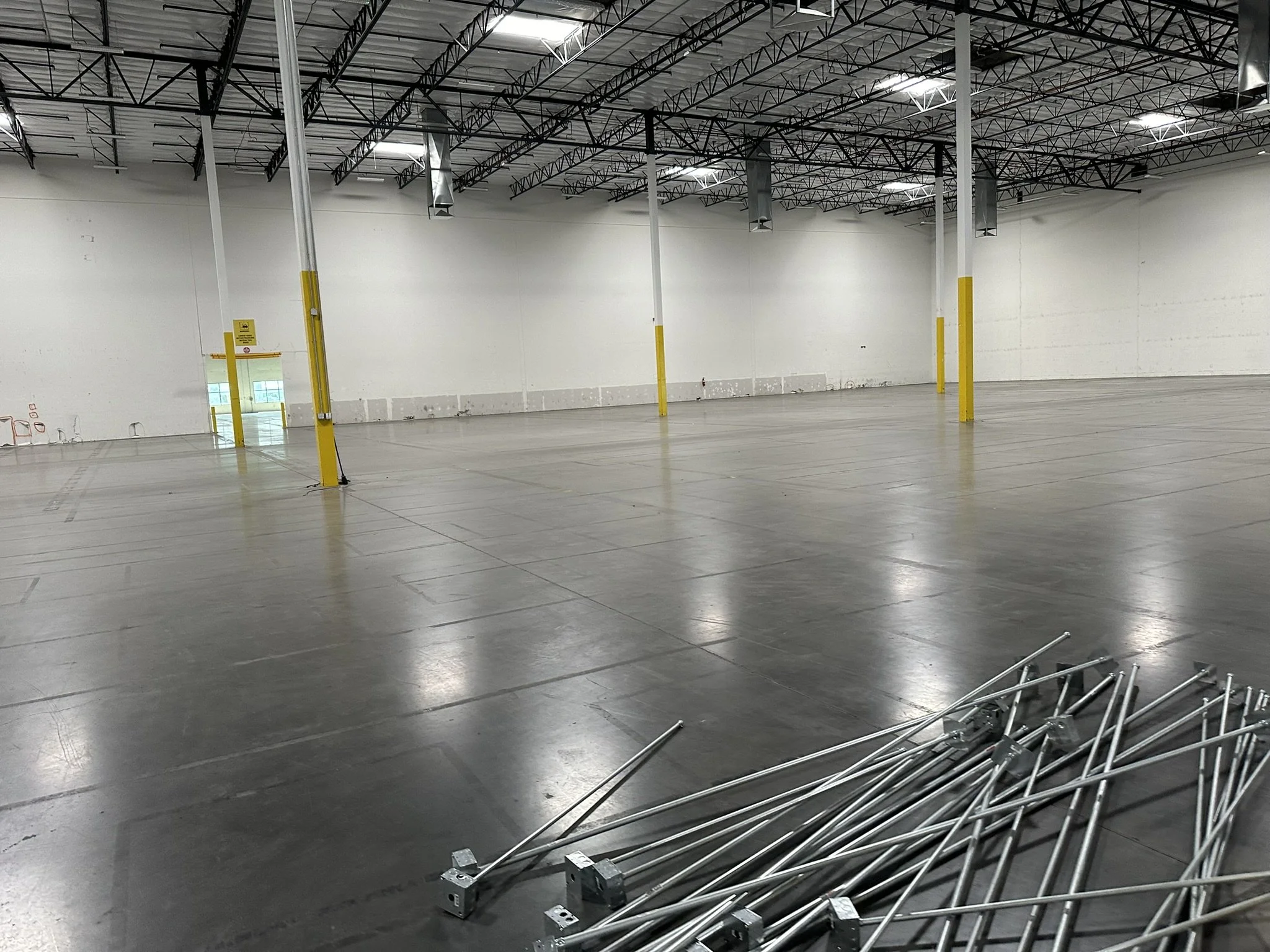 An empty warehouse or large store with a high ceiling, bright lighting, and a polished concrete floor. In the foreground, there are several metal shopping carts that are tipped over and scattered near the lower right corner. The walls are white and relatively plain, with some equipment hanging from the ceiling, and a small door visible on the left side.