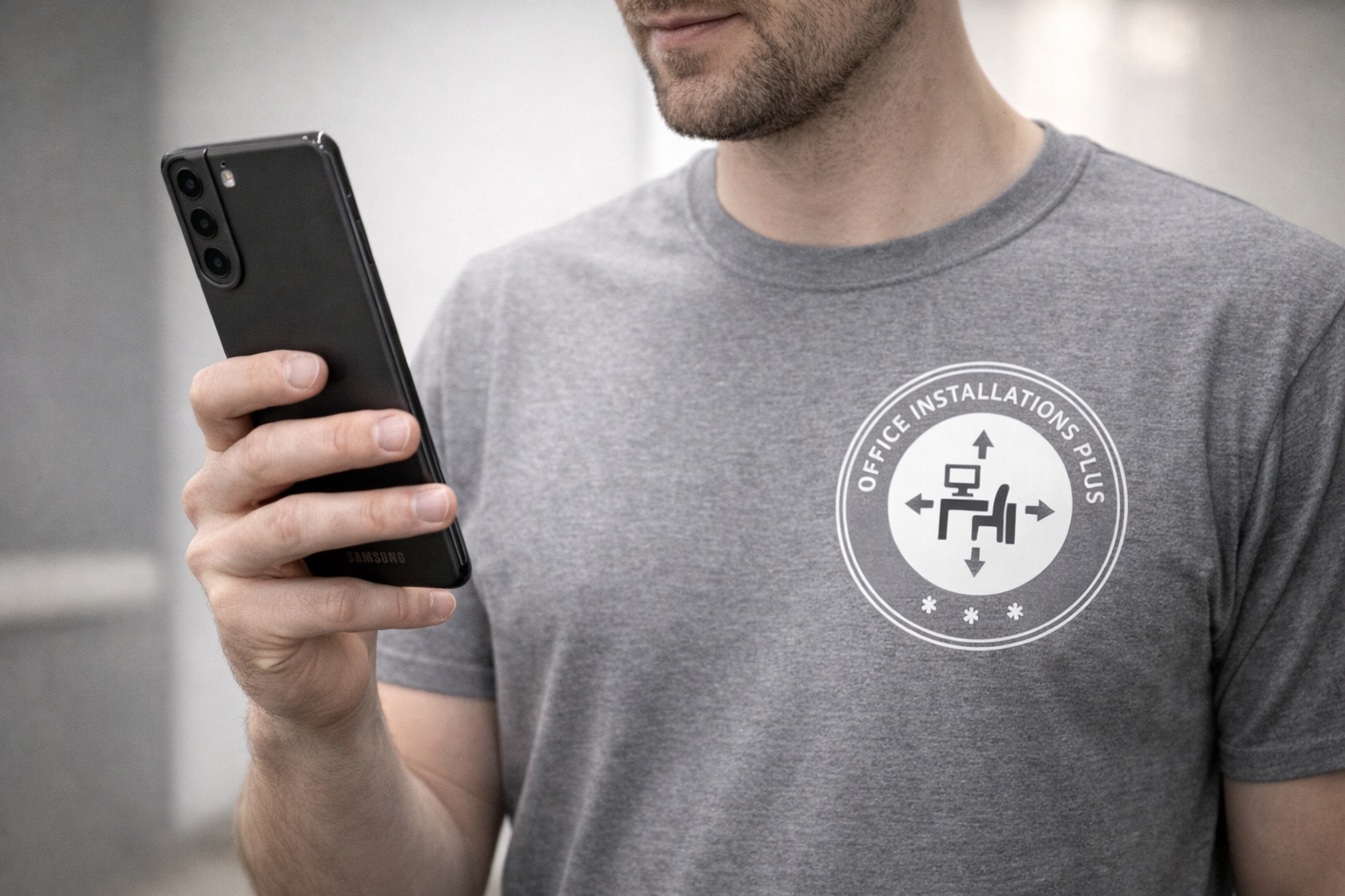 A man wearing a gray t-shirt with a circular logo that reads 'Office Installations Plus' and features a graphic of an office chair and arrows, holding a black smartphone symbolizing the attentiveness & responsiveness of Office Installations Plus.