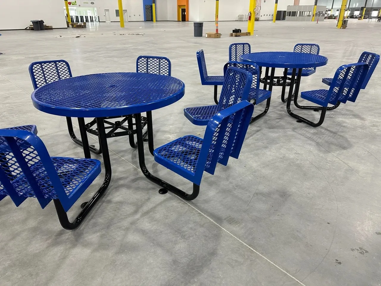 Two blue metal tables with matching blue metal chairs arranged in an empty warehouse or large indoor space with concrete floor.