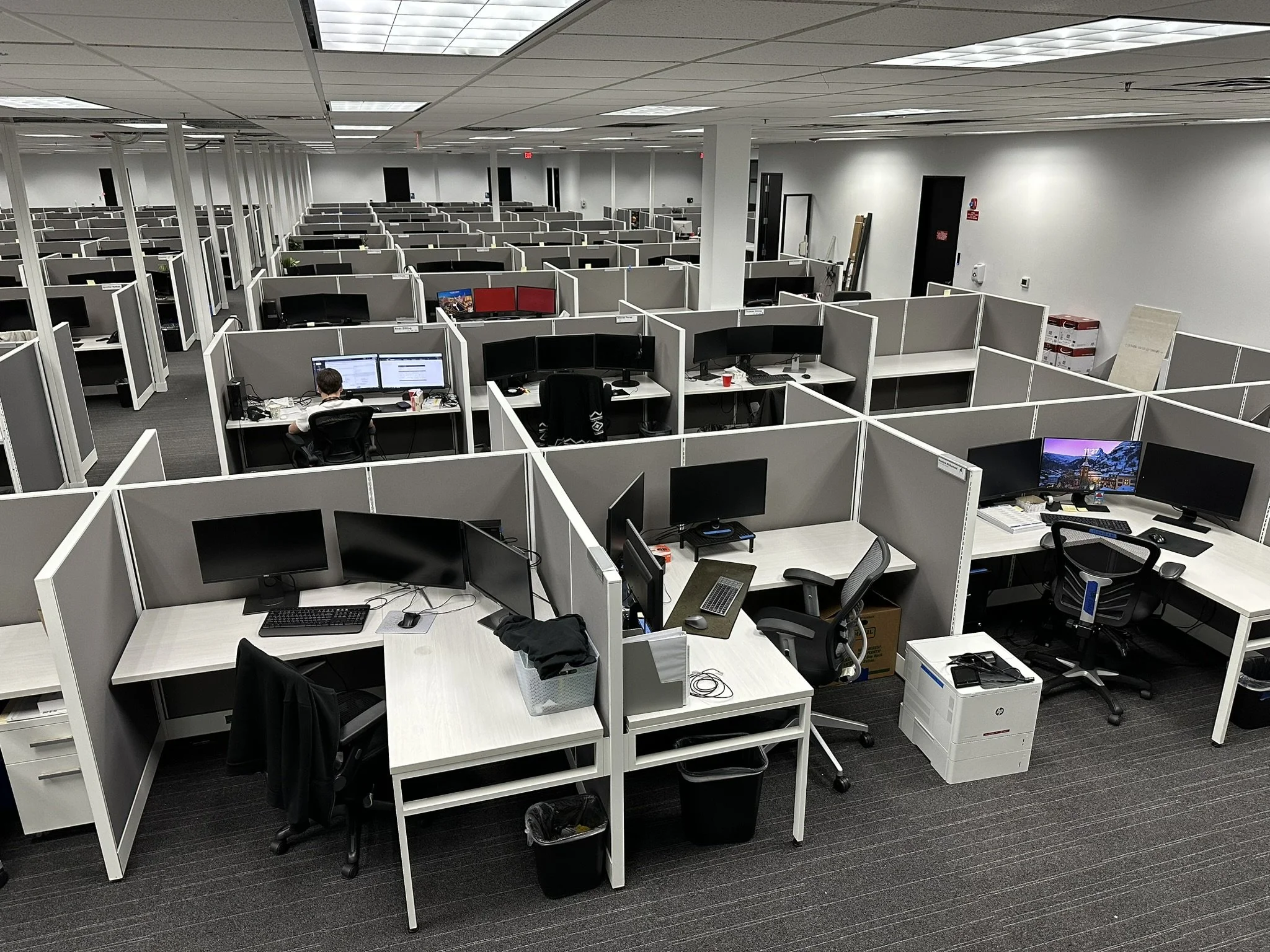 Empty office space with rows of white desks, each equipped with monitors, chairs, and adjustable lamps. Highlights professional office installation, workspace setup, furniture assembly, and modern commercial office build-out.