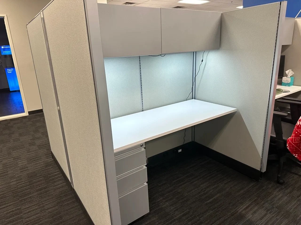 Empty office cubicle with a white desk, overhead storage, and a small filing cabinet, with a phone, tissue box, and computer on neighboring desks.