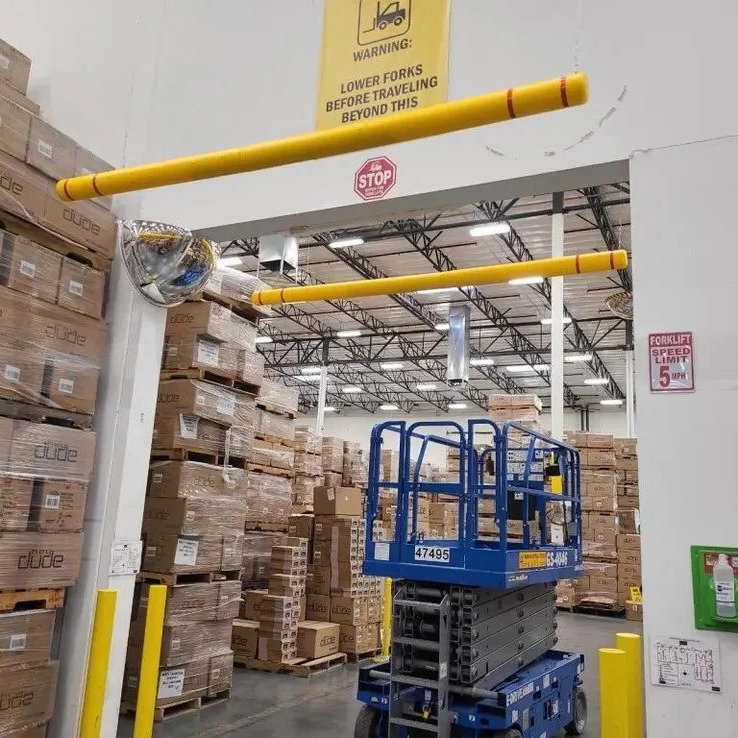 Warehouse with cardboard boxes on pallets, yellow protective barriers, a blue lift, and safety signs including a warning about lower forks and a stop sign.