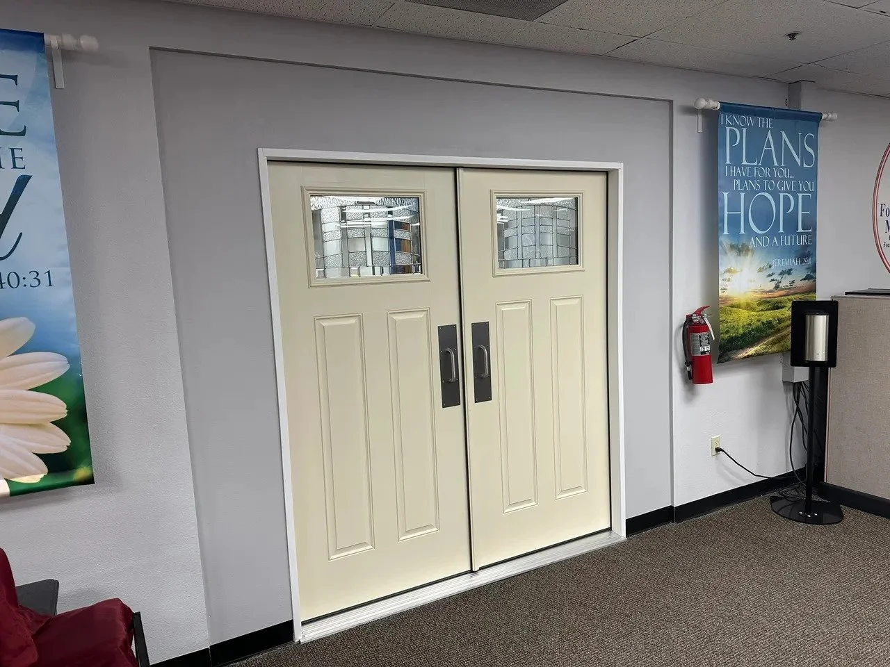 Double metal doors with small rectangular windows at the top, located in an indoor hallway with gray walls and carpeted floor. To the right is a fire extinguisher mounted on the wall, and a black stand with a fire alarm or speaker. On the walls are posters, one with a flower and the other with inspirational text and a landscape.