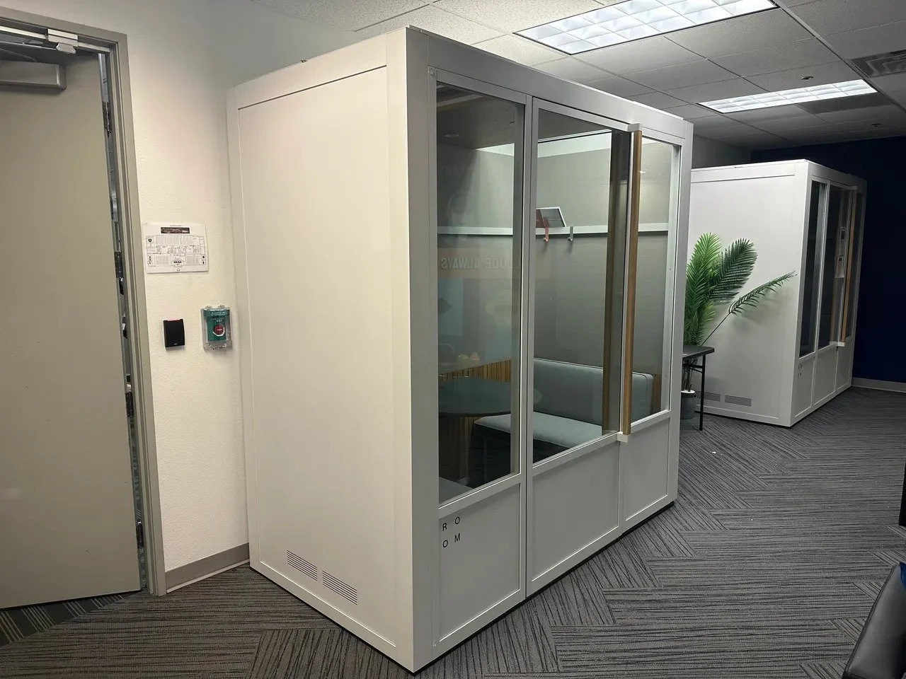 Office interior with white partitioned glass booths, a small table with a plant, and carpeted flooring.