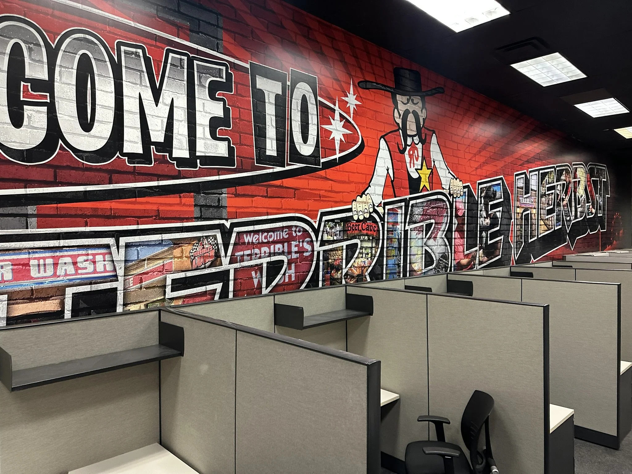 Empty office cubicle with a white desk, overhead storage, and a small filing cabinet, with a phone, tissue box, and computer on neighboring desks.