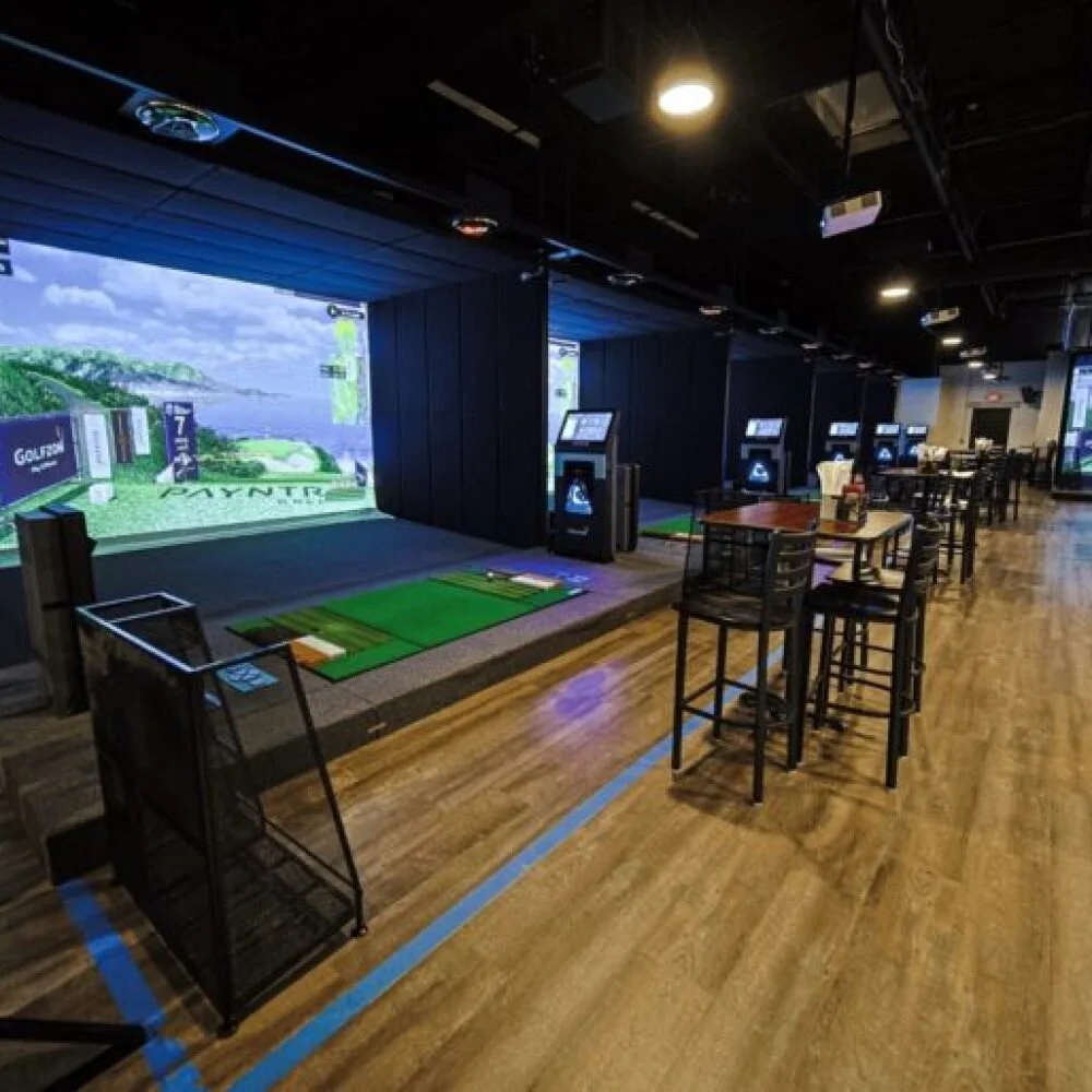 Indoor golf simulation room with multiple golf simulators, wooden floors, and seating area with tables and chairs.