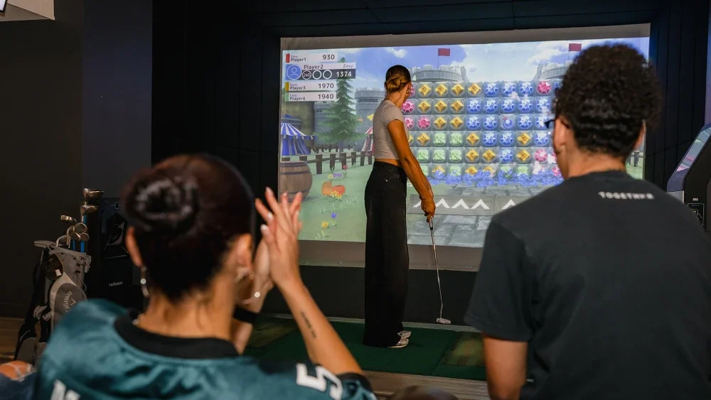 People watching a woman play an indoor golf arcade game on a screen