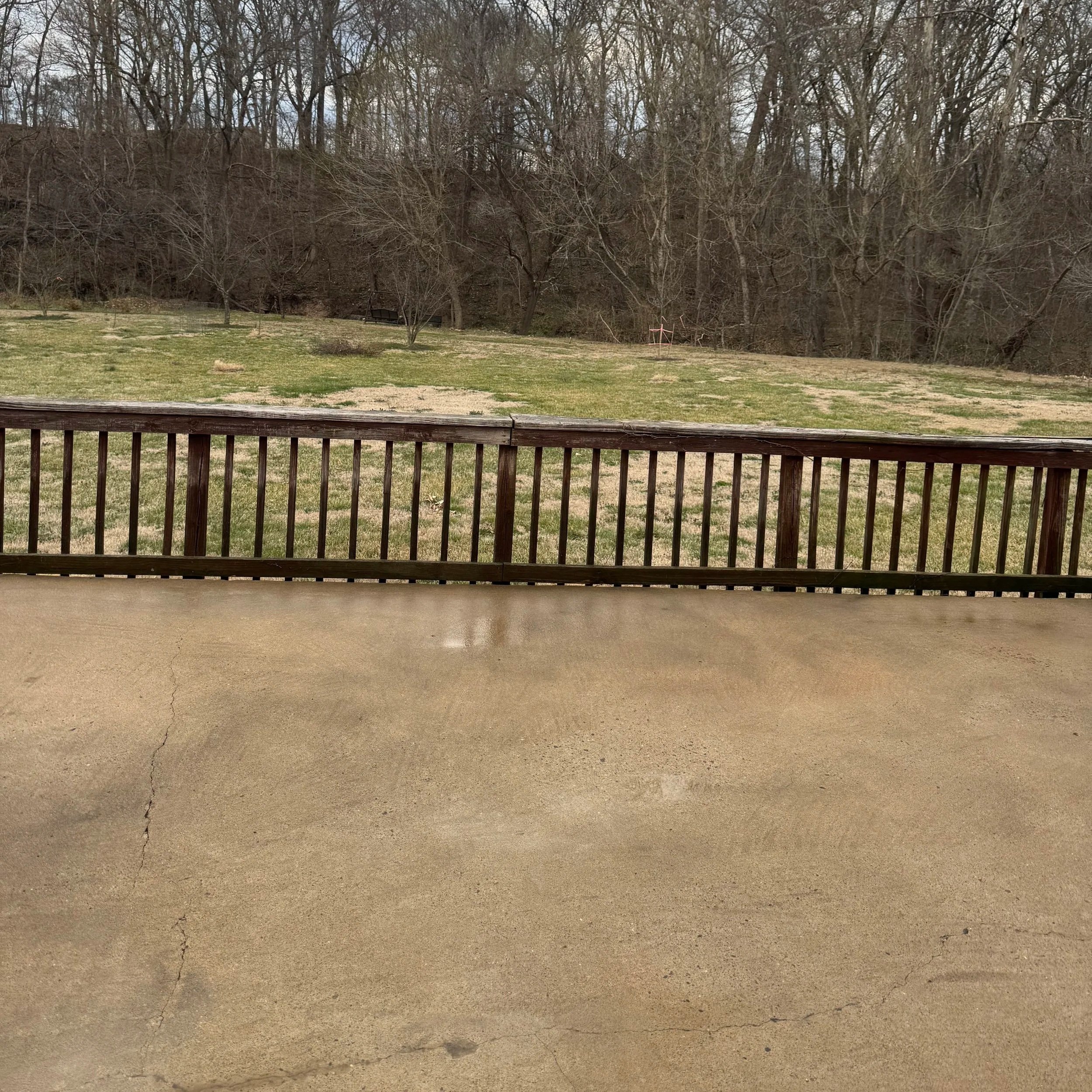 View from a concrete patio with a wooden railing, overlooking a grassy yard with patches of dirt and a wooded area in the background.