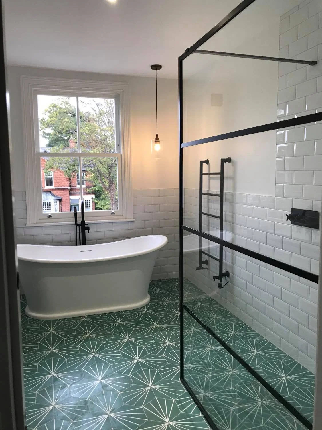 Modern bathroom with a freestanding bathtub near a window, a glass-enclosed shower with black fixtures, geometric patterned tile floor, and a hanging light bulb.