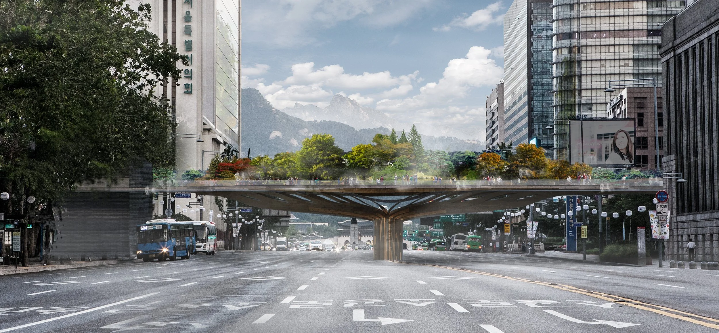 City street with a built-in park and pedestrian bridge overhead, surrounded by tall office buildings, trees, and mountains in the background.