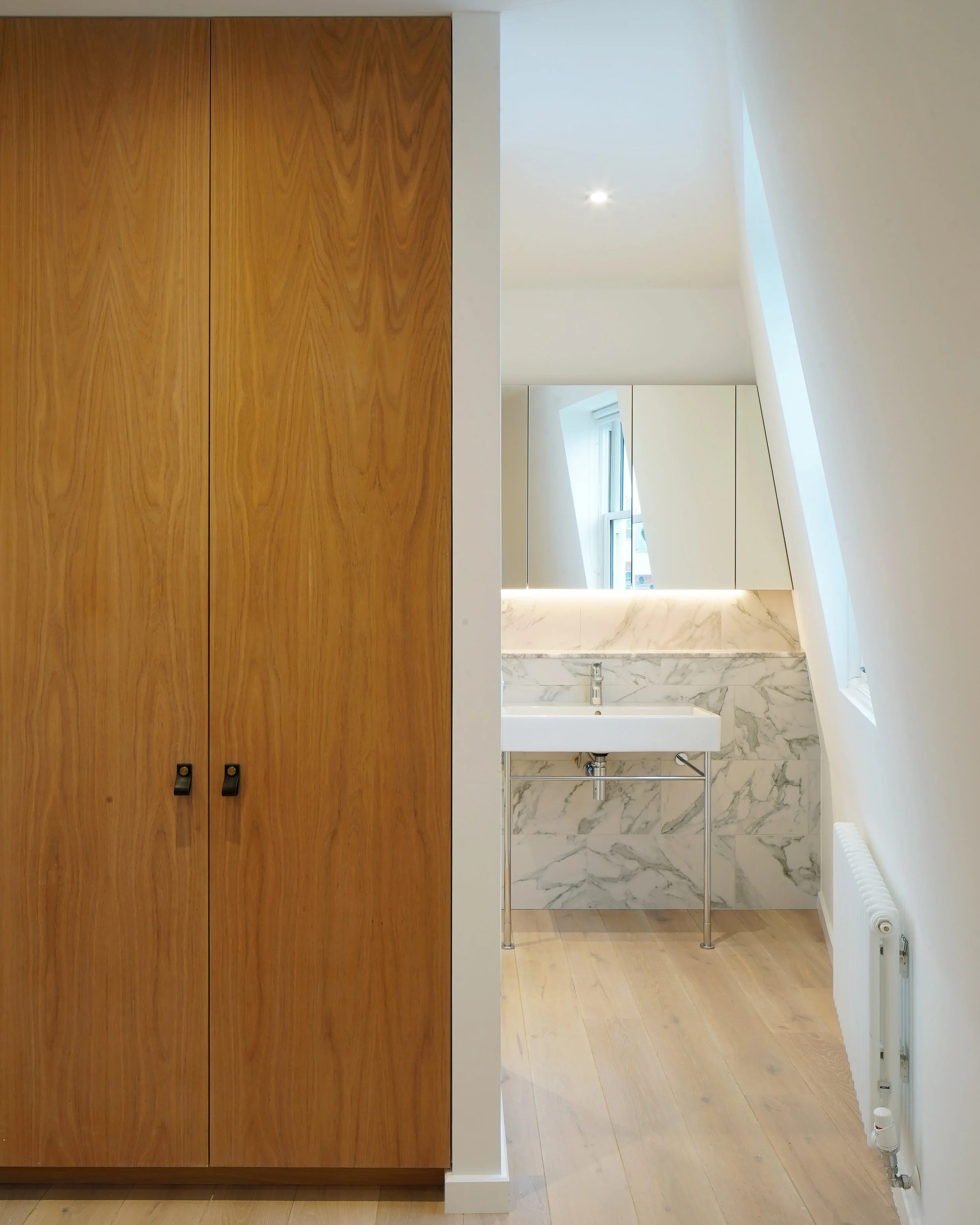 View of a modern bathroom with a white sink, marble backsplash, cabinets, a window, and a sloped ceiling, seen through an open space next to a wooden wardrobe.