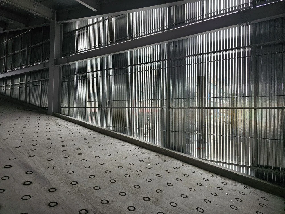 Underground pedestrian walkway with metal railing, transparent grills, and a textured rubber floor, with sunlight filtering through the grills.