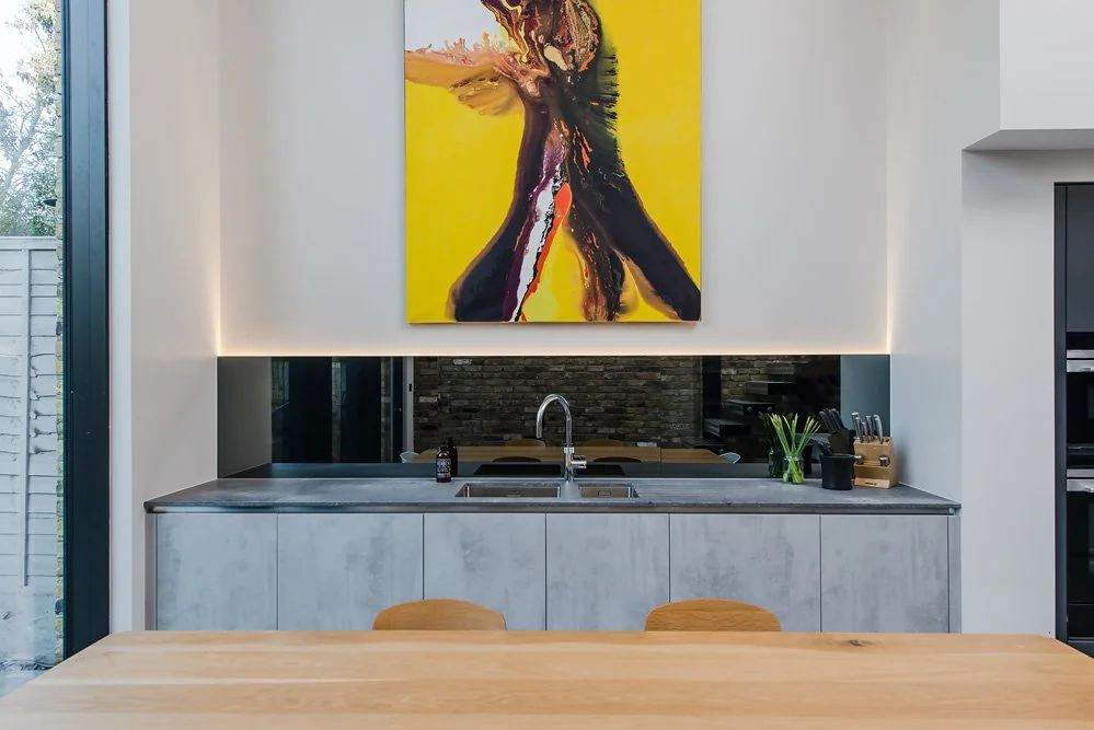 Modern kitchen with a concrete countertop, a sink, and a large yellow and black abstract painting on the wall. The kitchen has minimal decor with a potted plant and utensils. There is a large window to the left with a view of trees.