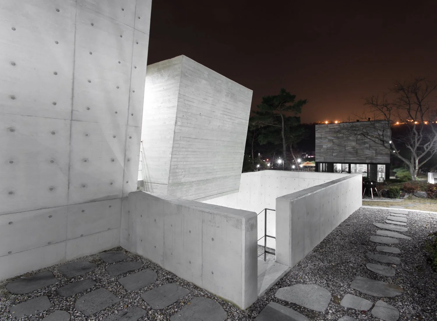 Nighttime view of modern concrete buildings with sharp angles and textured surfaces, surrounded by a stone pathway and sparse trees.