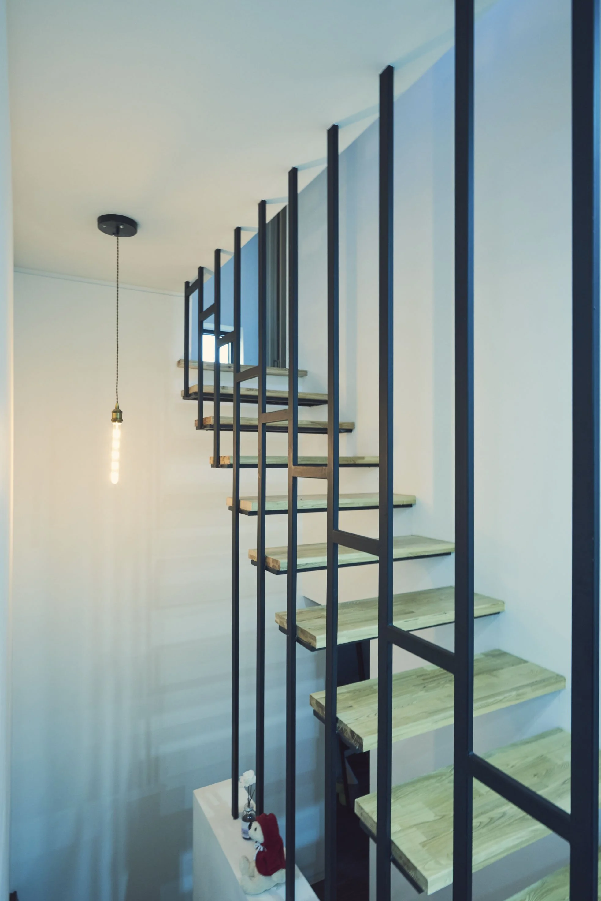 View of a modern staircase with wooden steps and black metal framing in a minimalistic home. A hanging light bulb is visible on the ceiling, and a small decorative item with a red scarf is on a white pedestal at the bottom of the staircase.