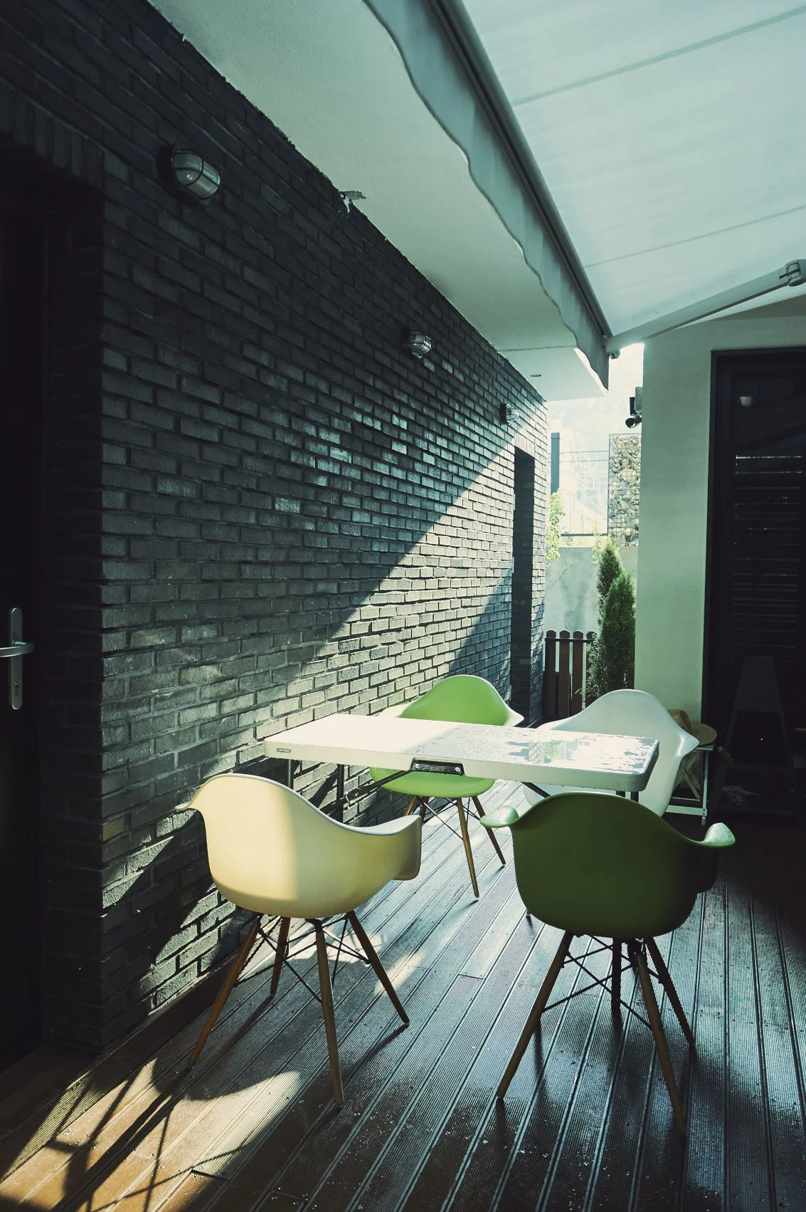 Outdoor patio with white table and four modern chairs, against a black brick wall, shaded by a white retractable awning, with green plants in the background.