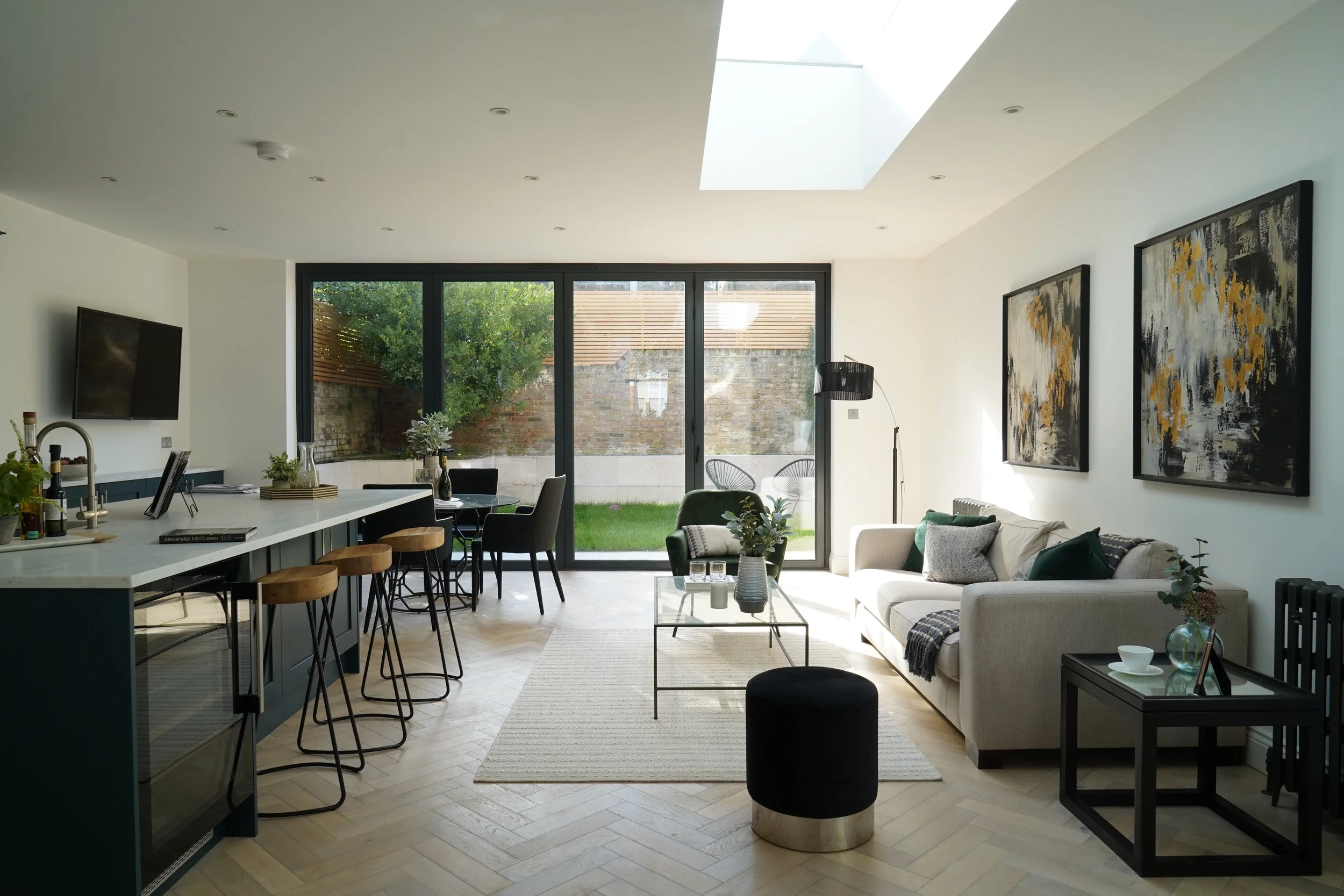 Modern living room with large glass sliding doors, light wood flooring, white couch, black side table, and wall art, with a skylight in the ceiling.