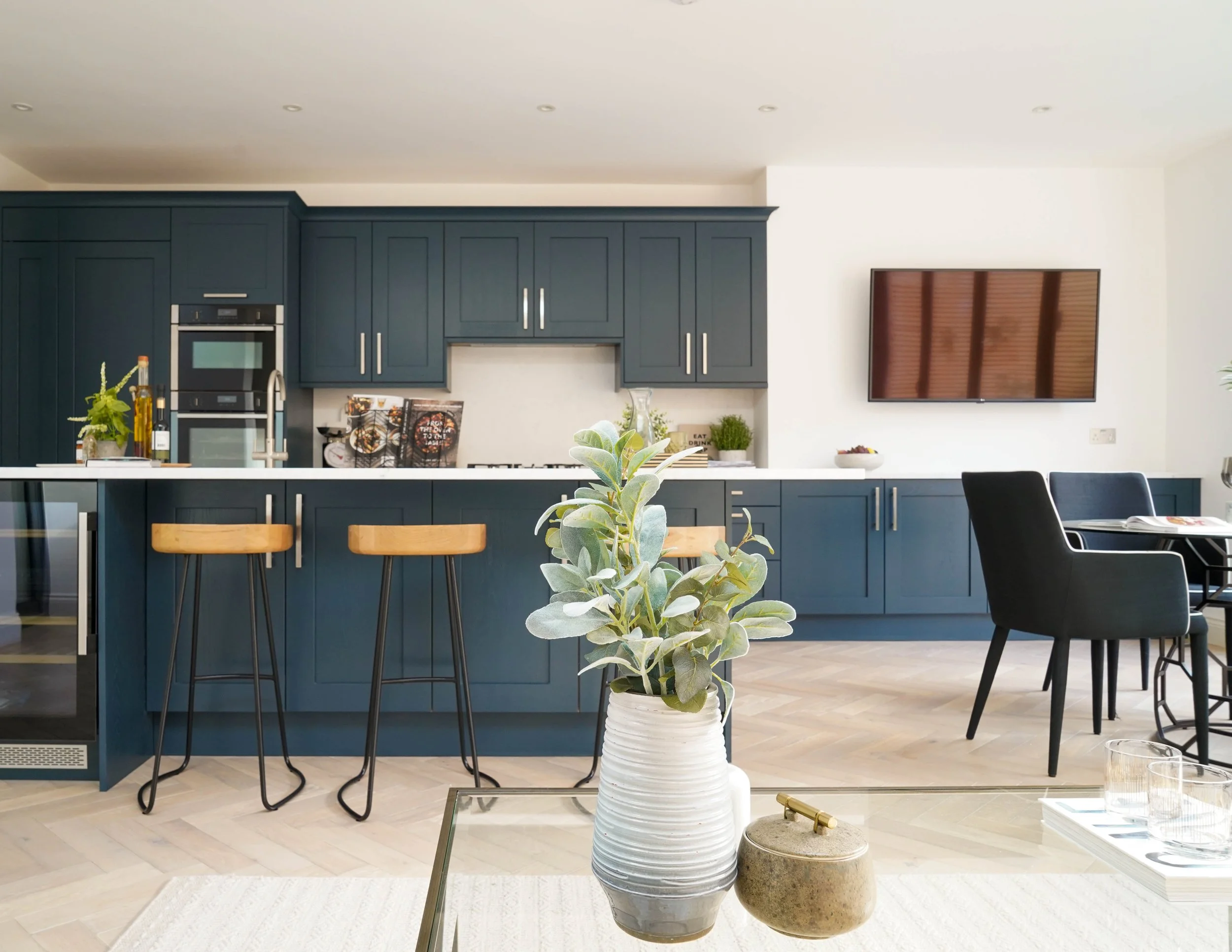 Modern kitchen with dark blue cabinets, white countertop, mounted flat-screen TV, bar stools, dining area with black chairs, potted plant on a glass table.
