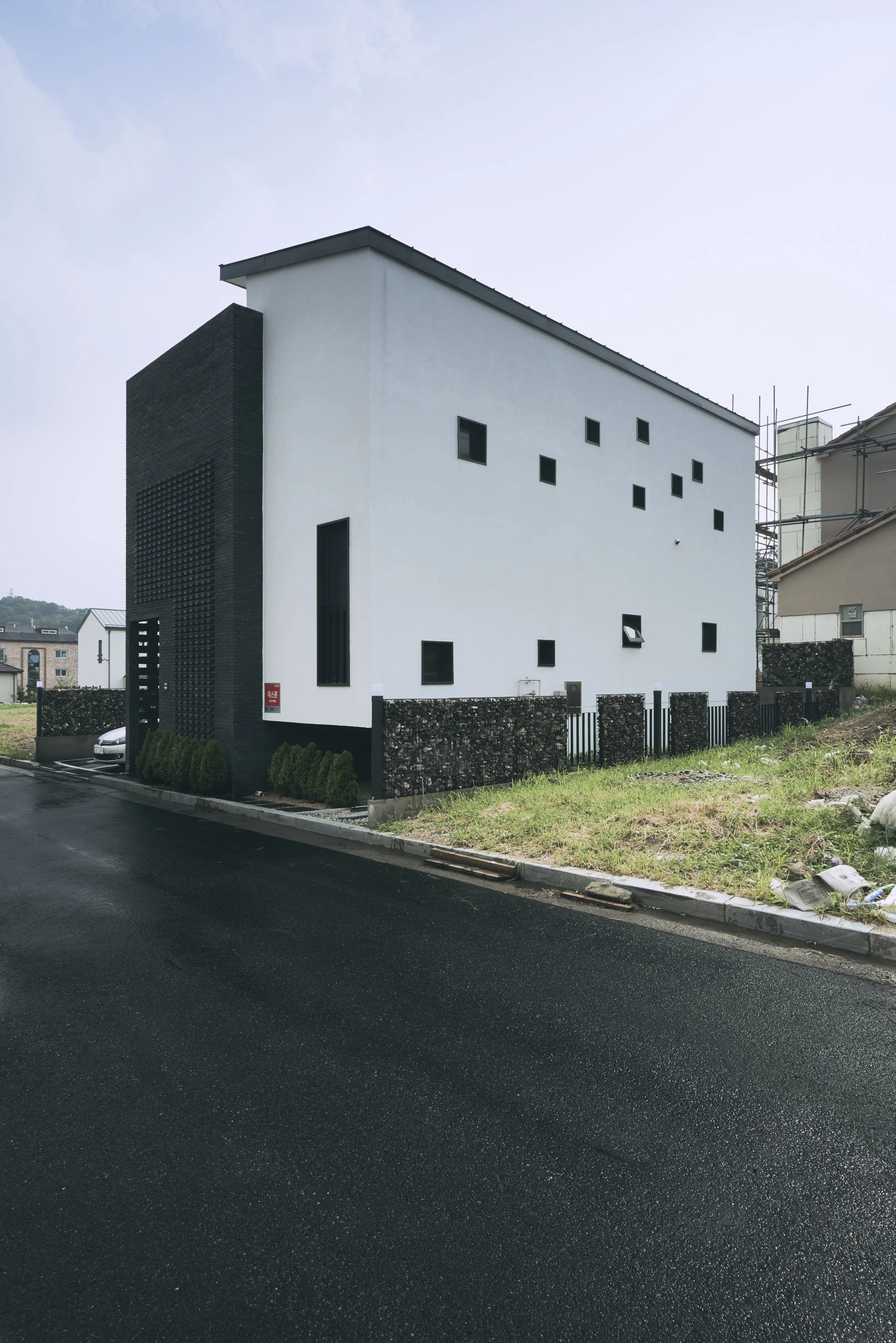 Modern white house with black accents and small square windows, located on a paved residential street, fenced yard with greenery, neighboring buildings, and scaffolding.