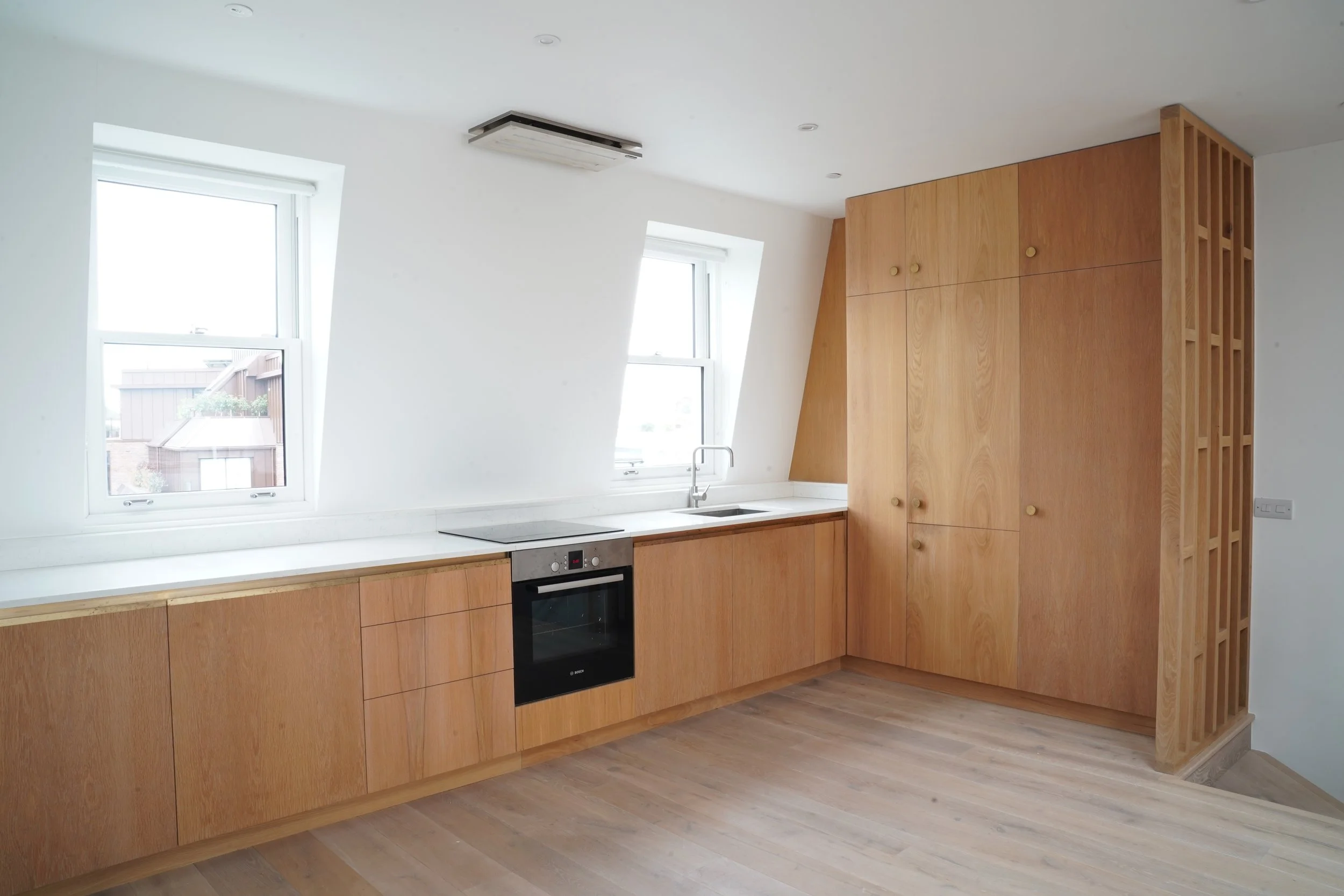 Modern kitchen with white walls, wooden cabinets, two windows, an oven, a sink, and a wooden partition at the end.