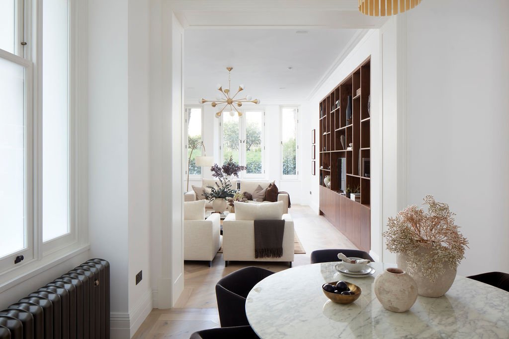A bright, modern living room and dining area with large windows, white walls, a round dining table with vases and bowls, and a built-in wooden bookshelf.