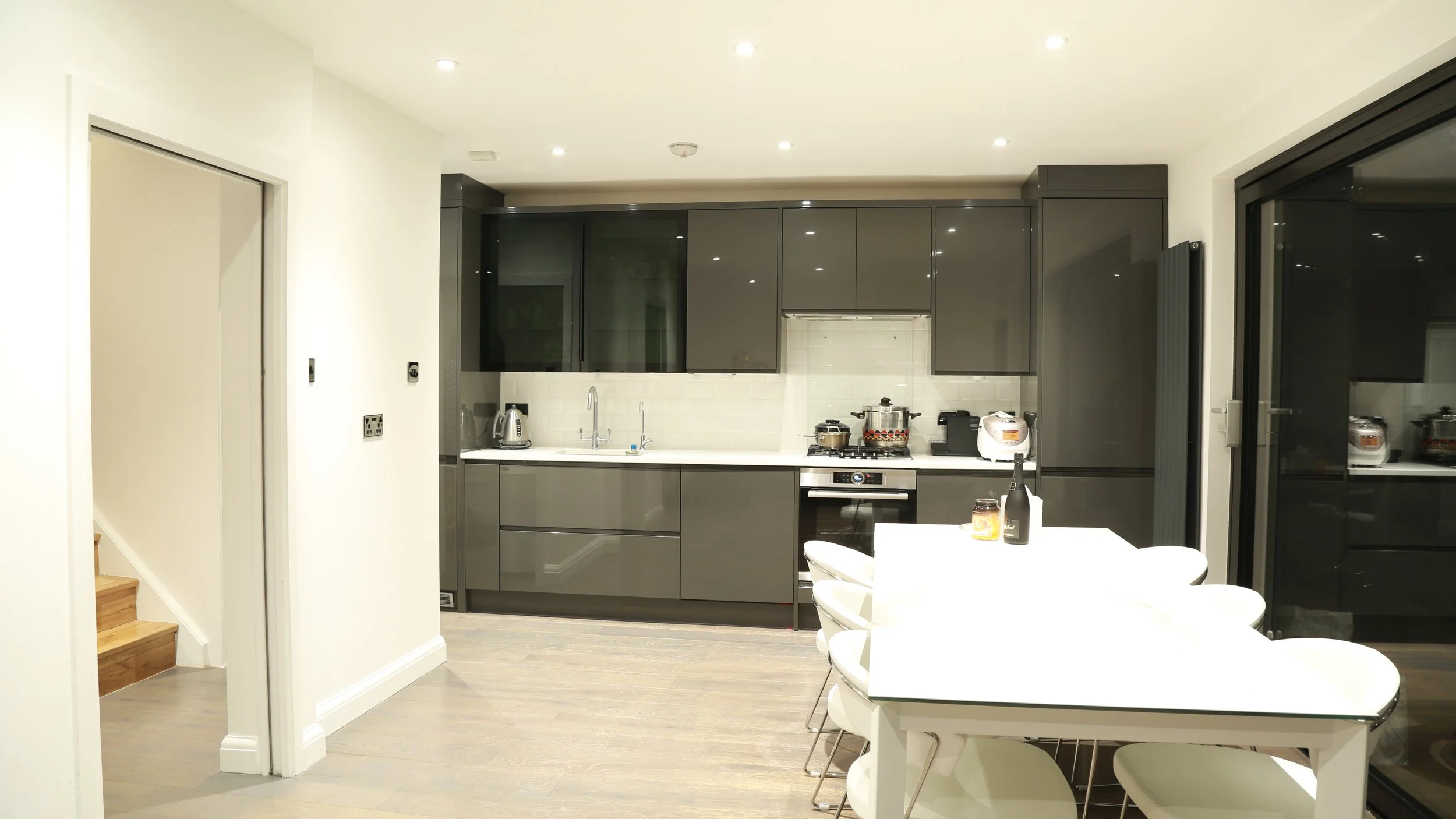 Modern kitchen with gray cabinets, white backsplash, and white countertop. Includes stove, oven, toaster, kettle, and various appliances. White dining table with chairs in the foreground, and sliding glass doors to the right.