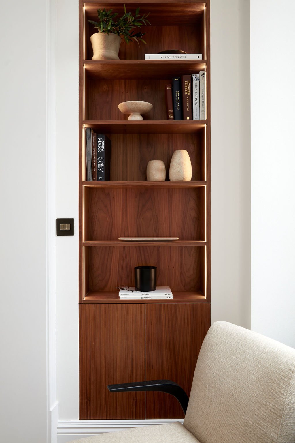 A tall wooden bookshelf with five shelves, decorated with books, vases, a plant, and a black container, positioned against a white wall.