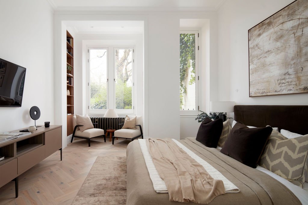 Modern bedroom with a large bed, window seat with two chairs, wall-mounted TV, and abstract artwork