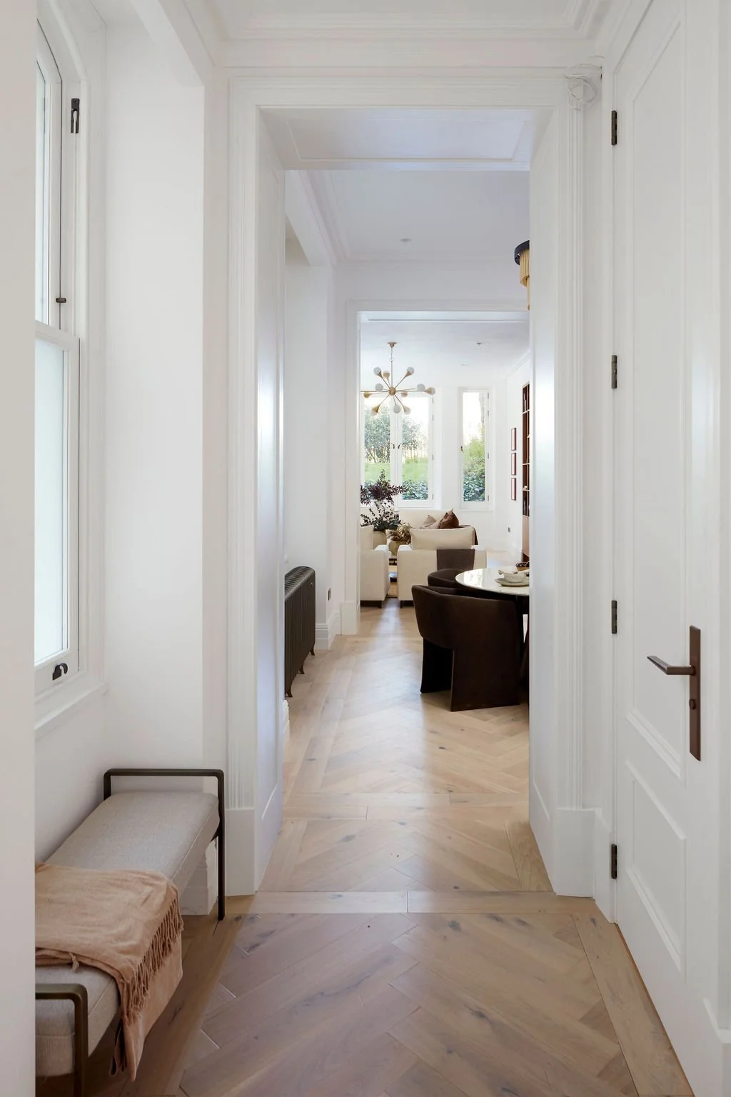 View through a bright, white-walled hallway into a sunlit living room with large windows, a chandelier, a seating area with cream and brown furniture, and hardwood floors.