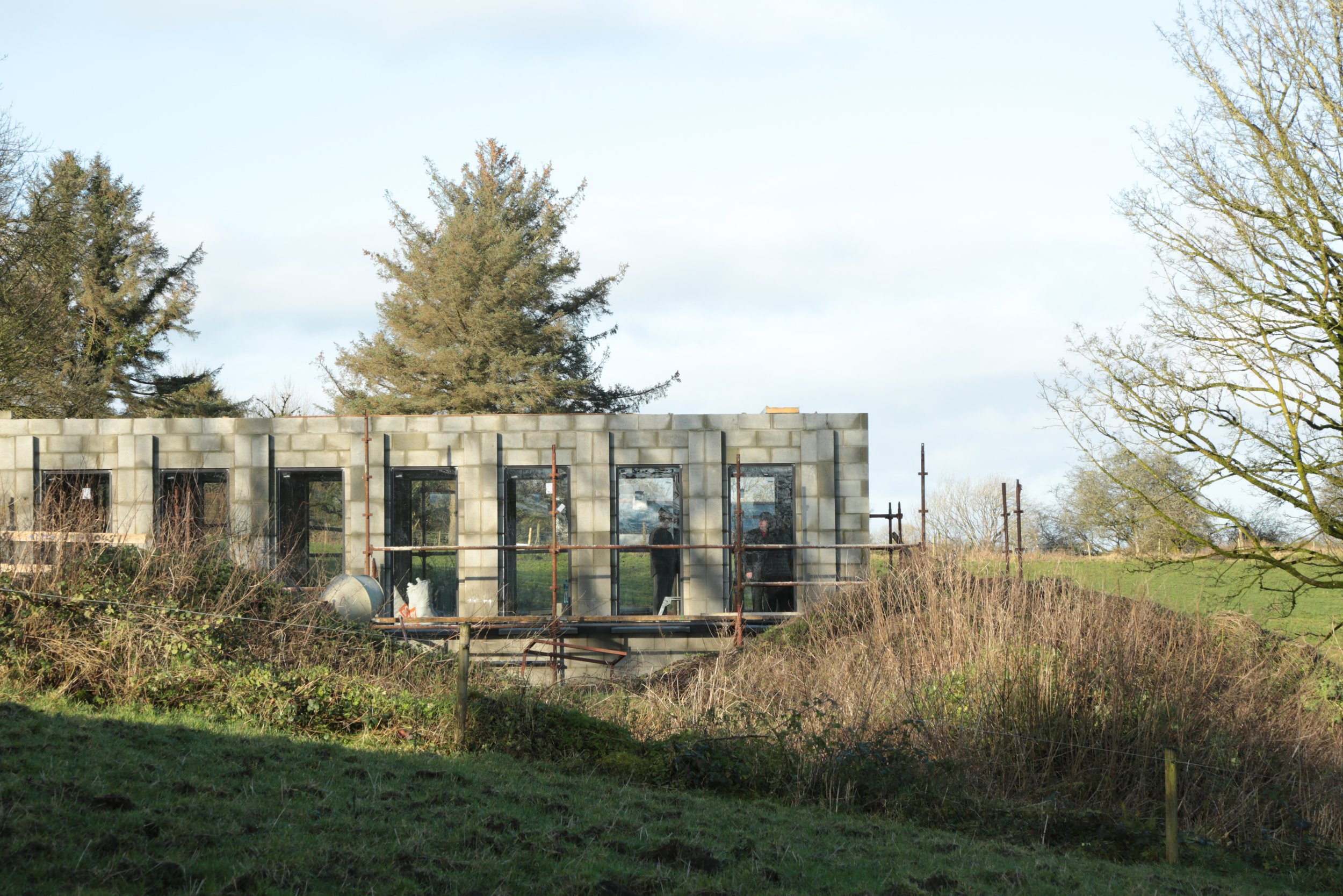 Construction site of a building with concrete blocks, scaffolding, and workers inside, surrounded by trees and grass.