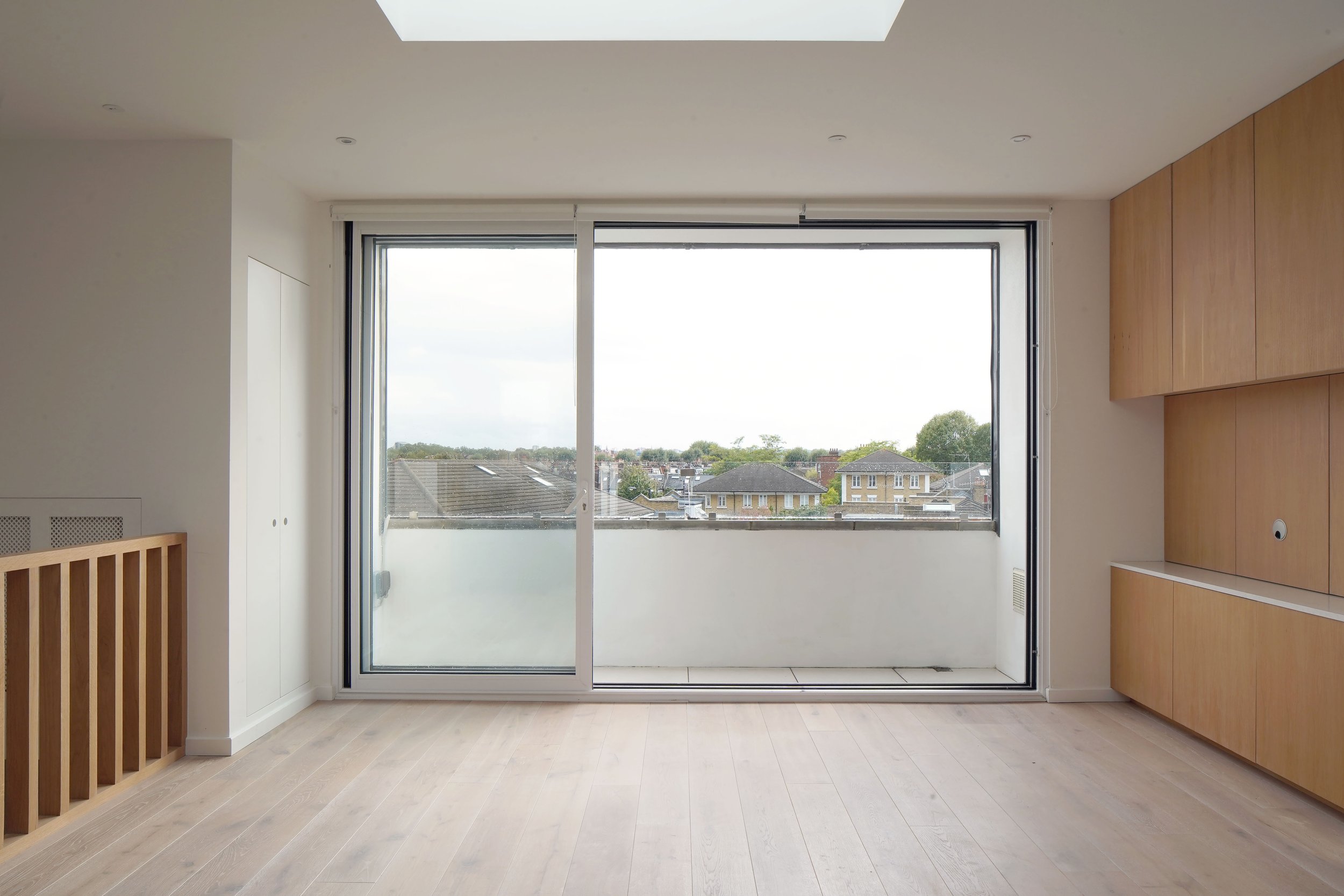 Empty room with large sliding glass door leading to a balcony, overlooking a neighborhood with houses and trees, light wood flooring, white walls, built-in wooden cabinetry on the right, and a wooden railing on the left.