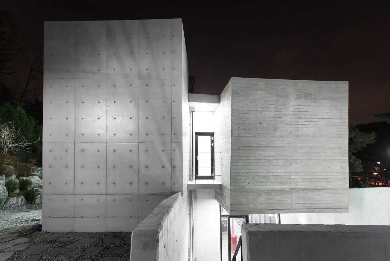 Night view of a modern concrete building with geometric shapes and small window, illuminated by exterior lighting.