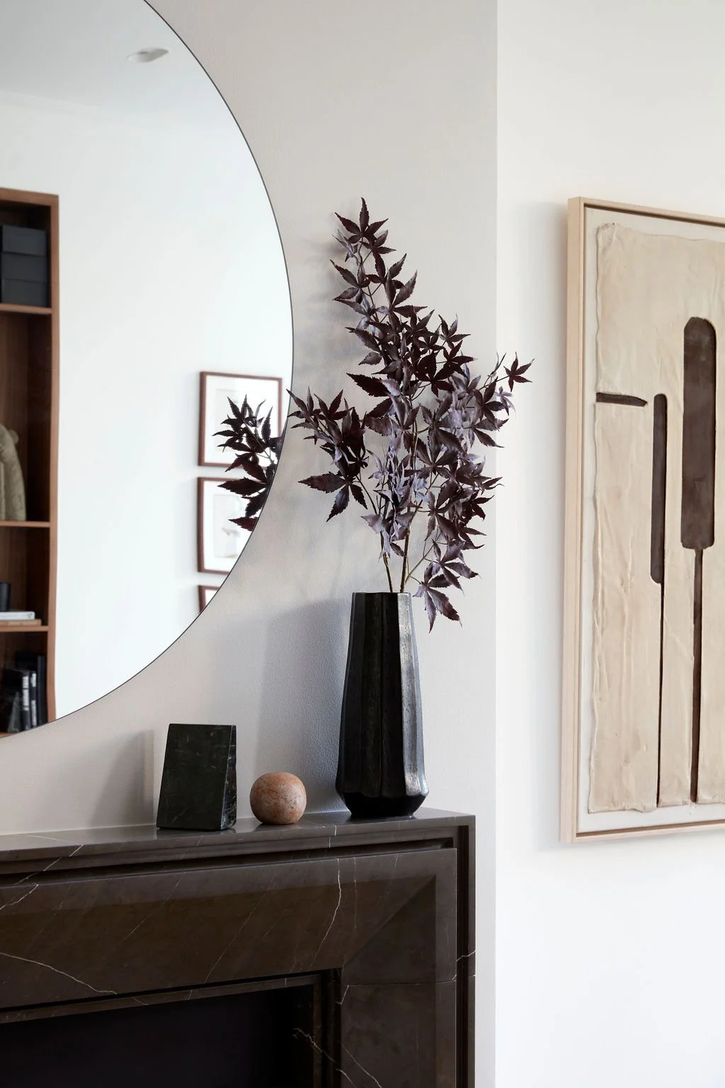 Decorative interior scene with a large round mirror reflecting part of the room, and a black vase with dark purple leaves on a dark marble sideboard. There are also two smaller decorative objects, a rectangular black object and a round stone sphere, 