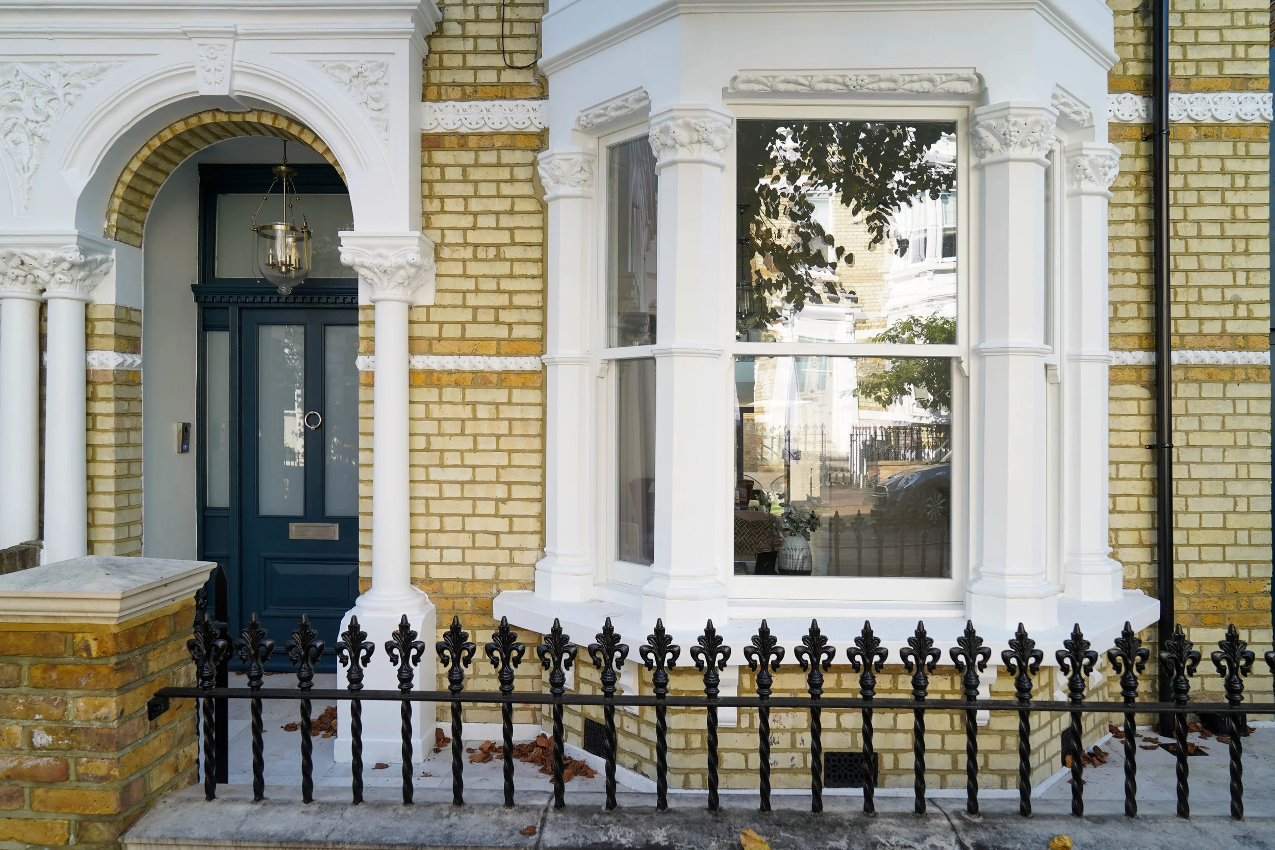 Exterior of a Victorian-style yellow brick house with white decorative trim and a blue front door, featuring a black iron fence in front.
