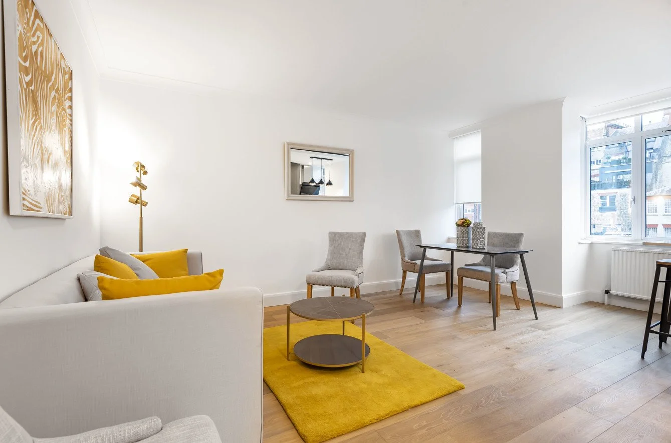 A bright living room with white walls and large windows, featuring a white sofa with yellow and gray pillows, a mustard yellow rug, a small round brown coffee table, two gray upholstered chairs, a black dining table with four chairs, a wall-mounted m