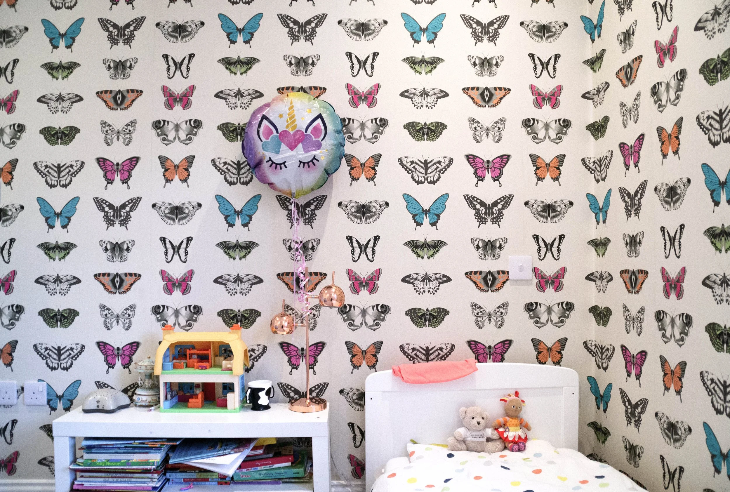 Children's bedroom with butterfly-themed wallpaper, a white bed with stuffed animals, a dollhouse on a white shelf, pink and copper balloons, and various books and toys.