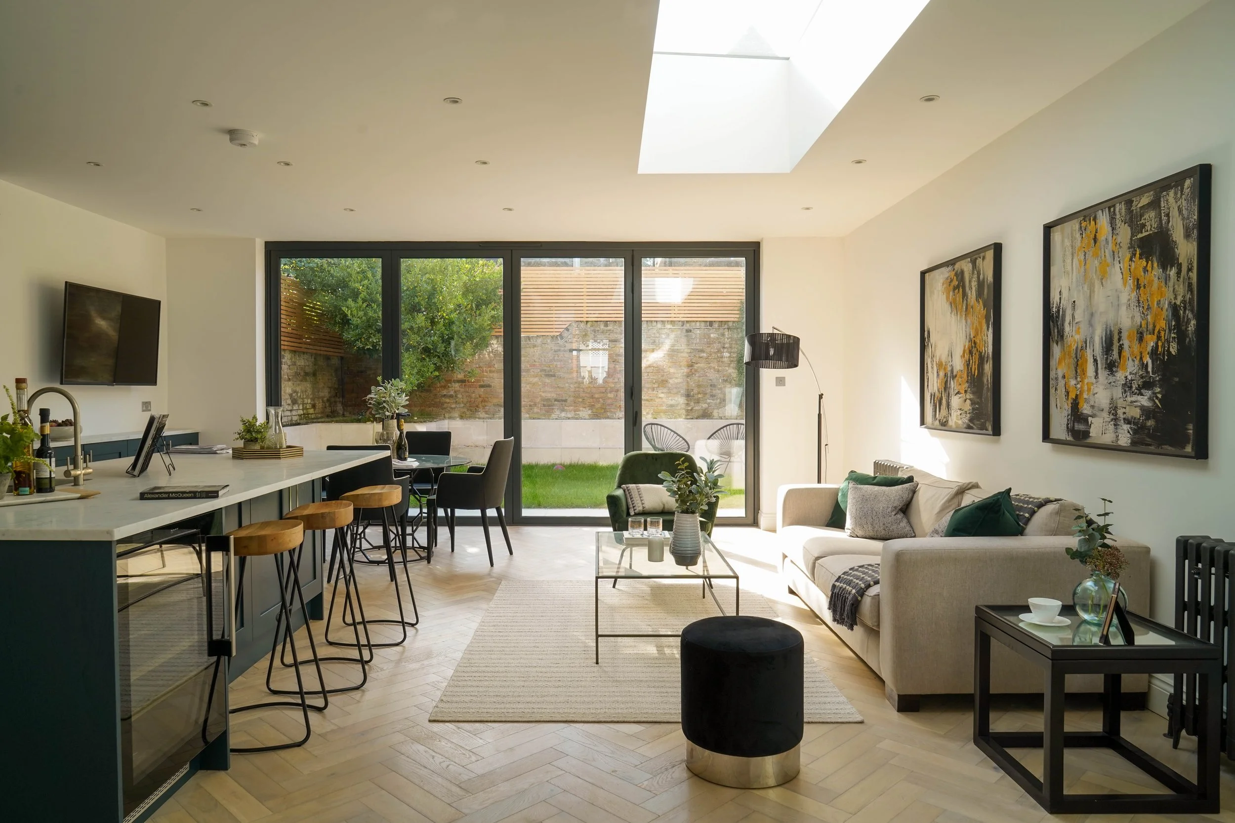 Modern living room with large glass sliding door leading to a backyard patio, featuring a white sofa, a black side table with drinks and decor, a green velvet armchair, a black floor lamp, artwork on the wall, a beige rug, and a kitchen island with s