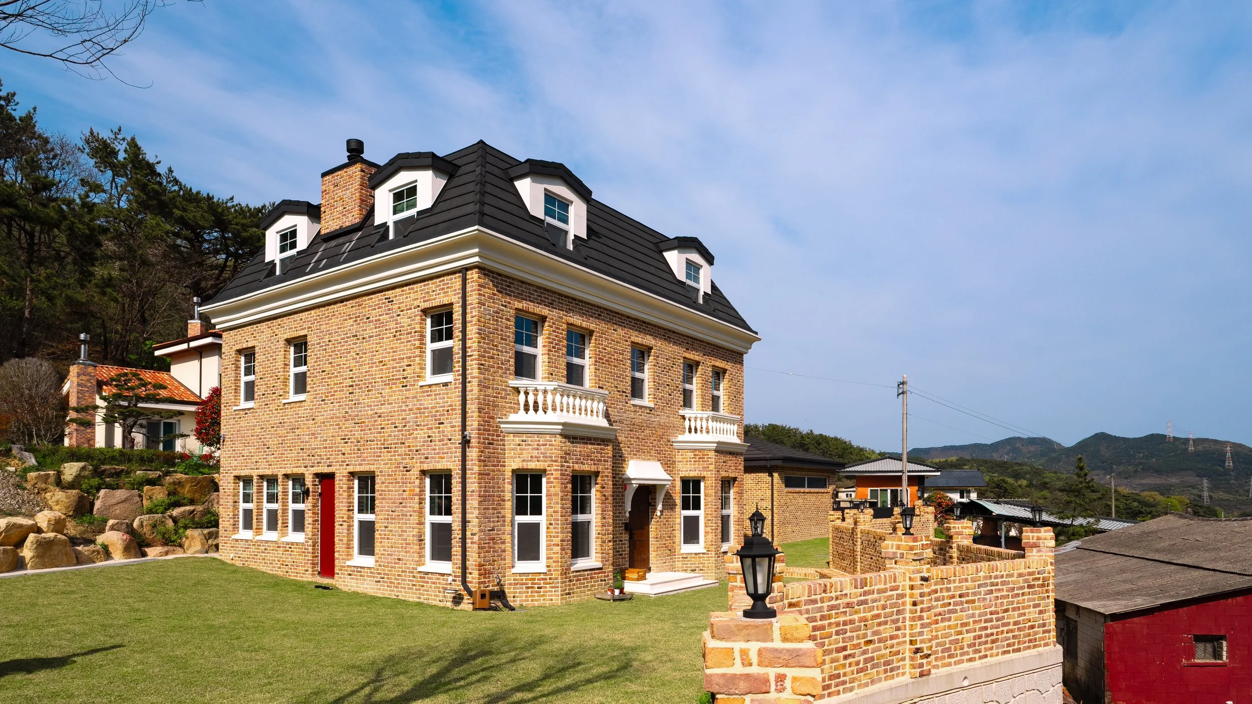 A large brick house with a black roof, multiple windows, and small balconies, surrounded by a well-maintained lawn and a brick fence with lantern-style lights.