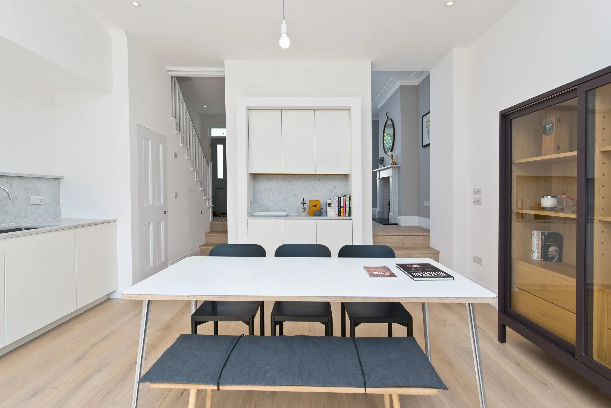 Modern dining room with white table, black chairs, a bench, and a glass display cabinet. Open kitchen with white cabinets and marble countertop visible on the left. Background shows a staircase, hallway, and fireplace.