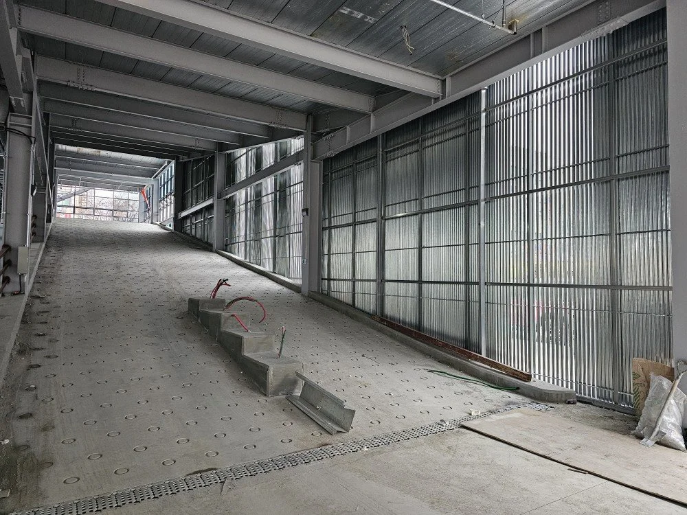 Underground parking ramp under construction with metal wall fencing, concrete surface, and some construction materials and pipes.