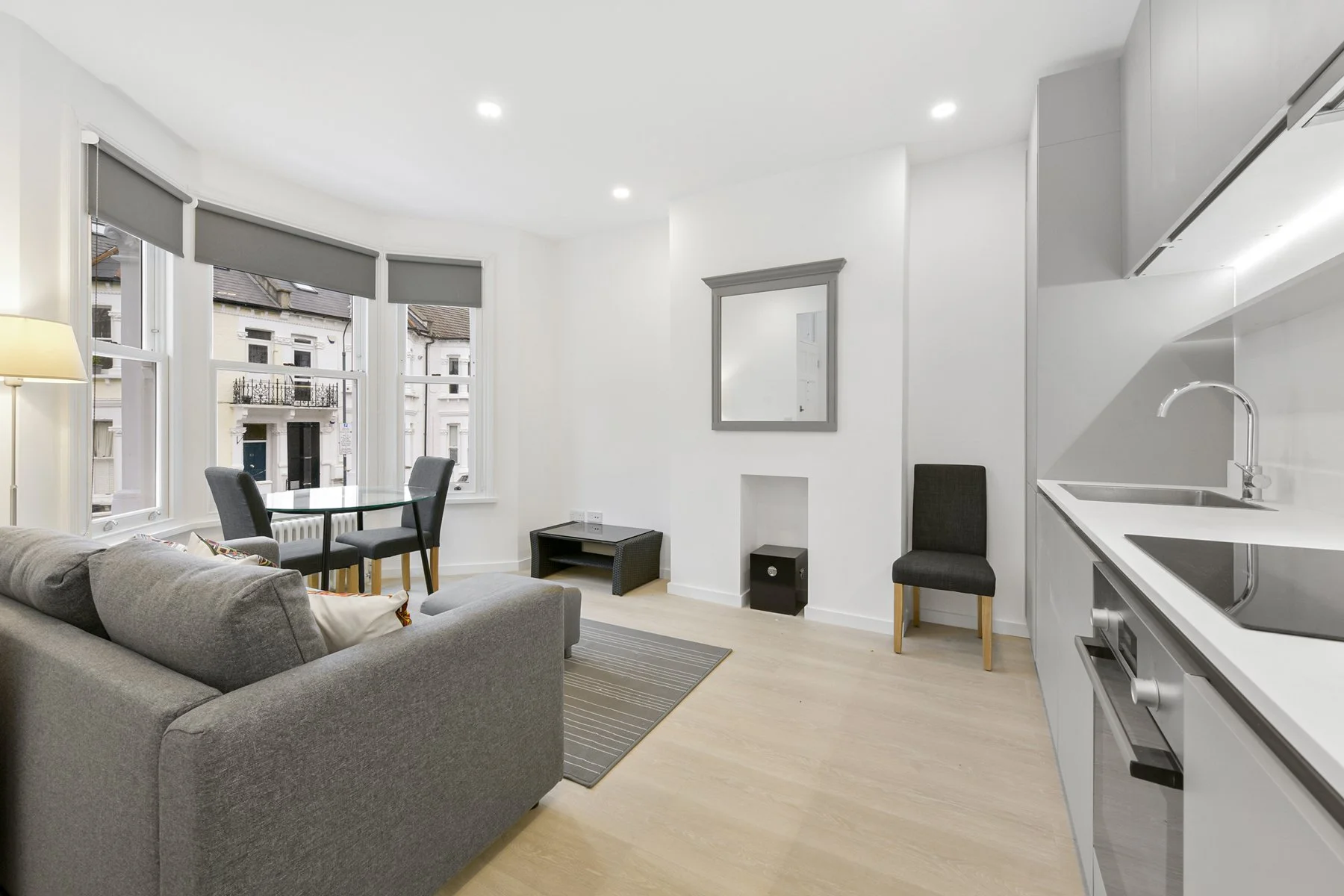 Open-plan living room and kitchen with grey couch, black dining chairs, windows with blinds, mirror on white wall, small black table, and kitchen sink