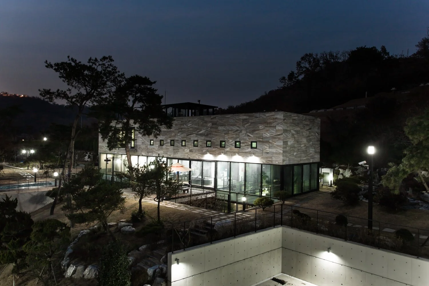 A modern building at night with large glass windows, surrounded by trees and outdoor lights, situated in a hilly landscape.