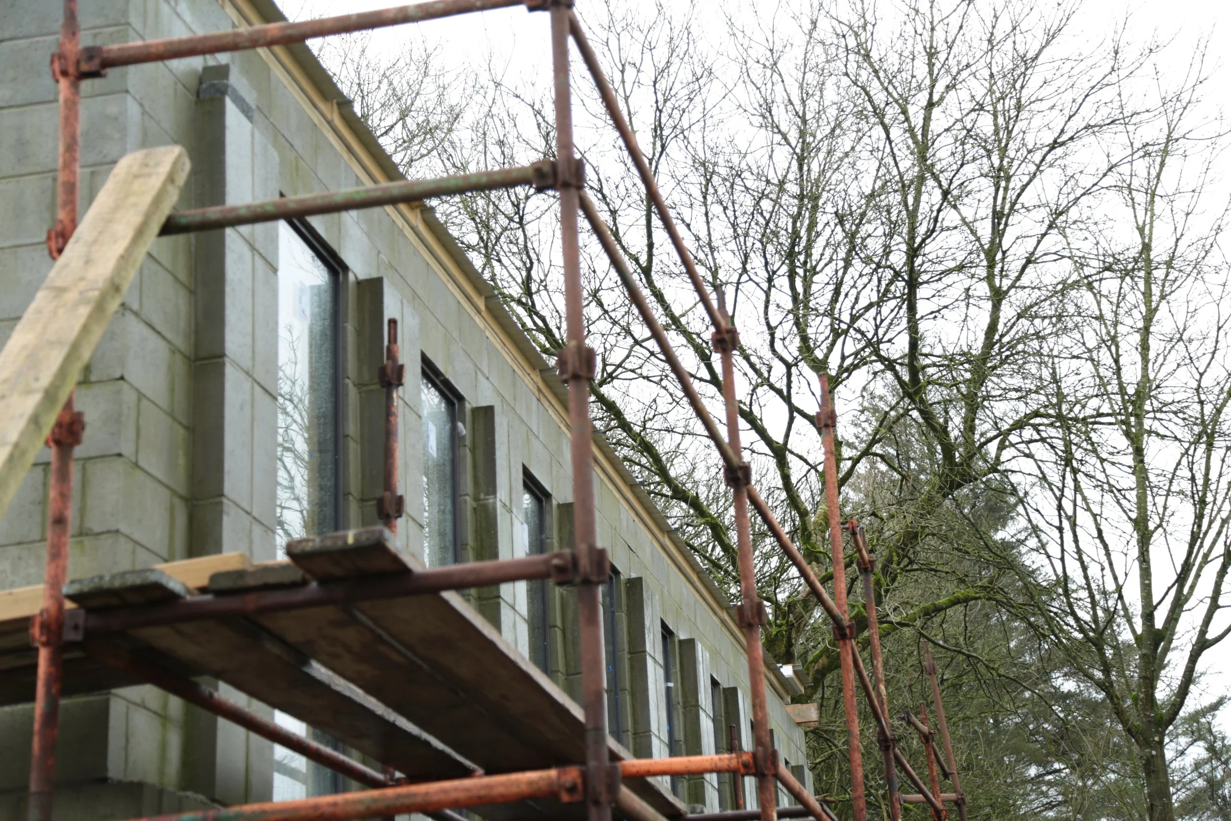 Construction scaffolding set up in front of a gray stone building with multiple windows, surrounded by leafless trees.